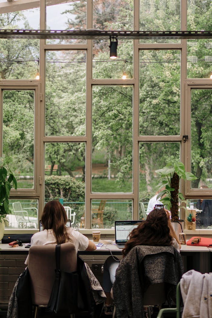Two women work on laptops in a cozy space with garden views through large windows.
