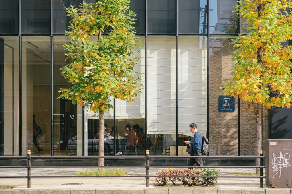Man walks past a modern glass building facade with trees and pedestrians inside.